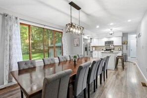 A modern kitchen with a central island, large windows, and a dining area with a wooden table and grey upholstered chairs. - The Birch: Poconos Getaway • Hot Tub, Game Room, Fire Pit • Sleeps 10 (Blakeslee)