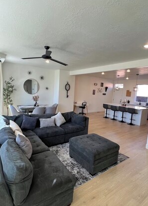 A modern living room with a sectional sofa, ottoman, and a dining area with a kitchen in the background. - Gorgeous 3-bedroom condo in charming Cedar City perfect for relaxing (Cedar City)