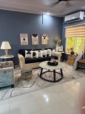A living room with a sofa, a round coffee table, a lamp, and artwork on the wall. - Charming 3-bedroom 2BTH house in Drax Hall with solar WiFi beach access pool (St. Ann Parish)