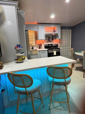 A modern kitchen with a central island, two bar stools, and a dining area with a table and chairs. - Charming 3-bedroom 2BTH house in Drax Hall with solar WiFi beach access pool (St. Ann Parish)