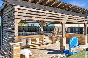 An outdoor kitchen area with a grill, sink, and storage cabinets, surrounded by a wooden pergola and a pool with a blue chair.