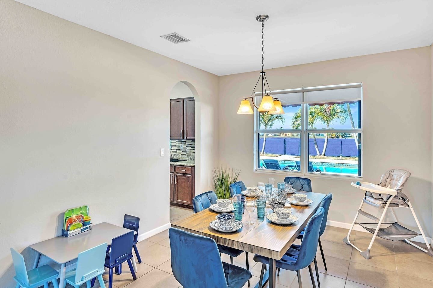 A dining area with a table set for four, a high chair, and a view of a swimming pool through large windows.