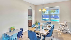 Comedor con una mesa dispuesta para cuatro personas, una trona y vista a una piscina a través de amplias ventanas.