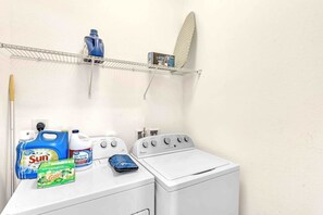 A laundry area with a washing machine, detergent bottles, and a shelf with a surfboard.