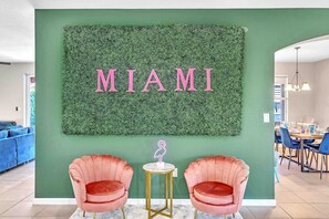 A room with a green wall, a large 'MIAMI' sign, two pink chairs, and a small table with a neon sign.