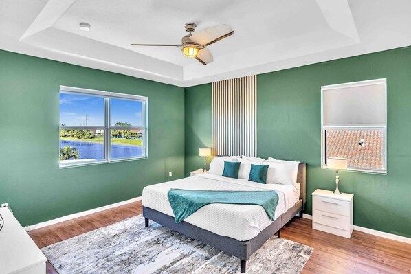 A bedroom with a bed, a nightstand, a lamp, a ceiling fan, and a window with a view of a body of water.