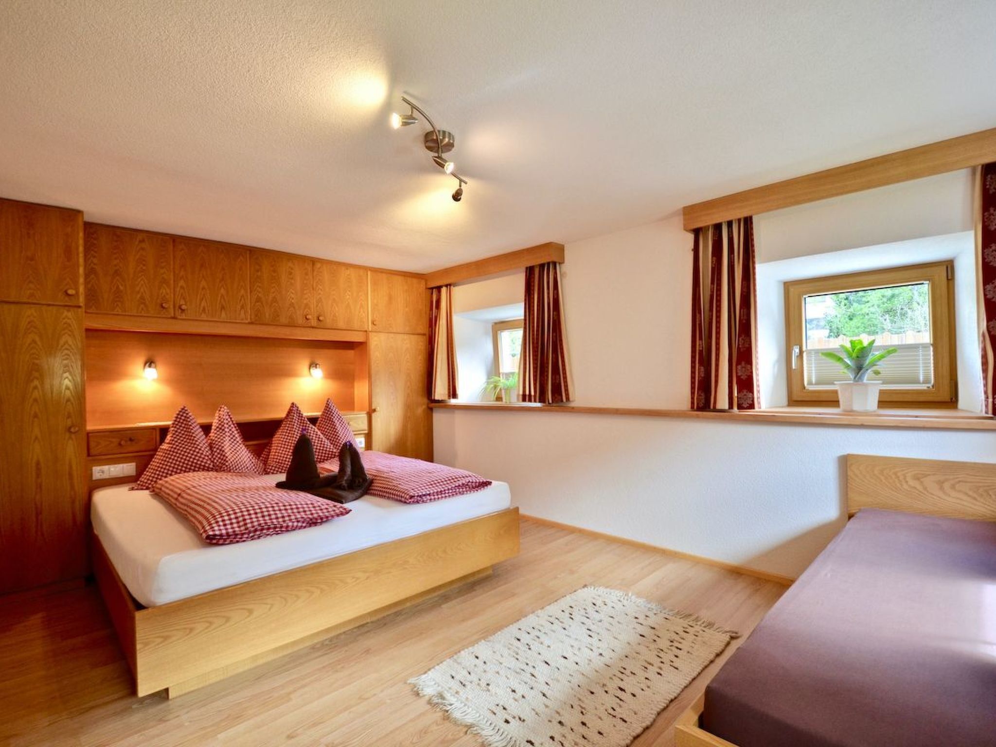 Ein Schlafzimmer mit einem hölzernen Bett, einem Nachttisch, einer Fensterbank, Vorhängen und einer Deckenleuchte.