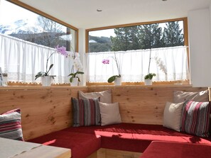 A cozy seating area with a wooden bench and cushions, large windows with curtains, and potted orchids on the windowsill. - Chalet Appartement Hörndle in Umhausen by Interhome (Niederthai)