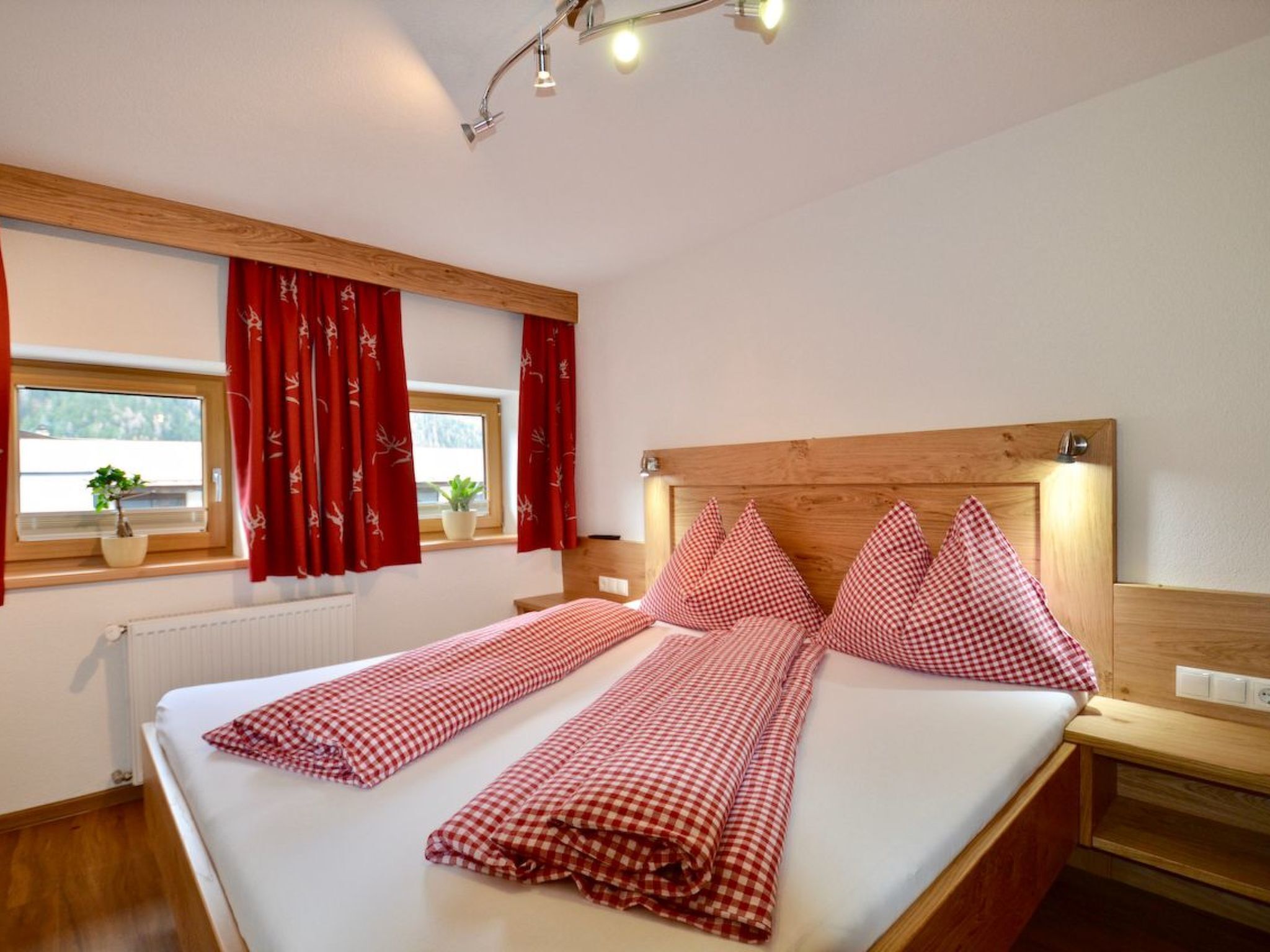 A hotel room with a large bed, red checkered bedding, wooden headboard, a window with red curtains, and a potted plant on the windowsill.