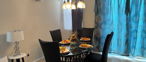 A dining area with a round glass table, black chairs, a chandelier, and a window with patterned curtains.