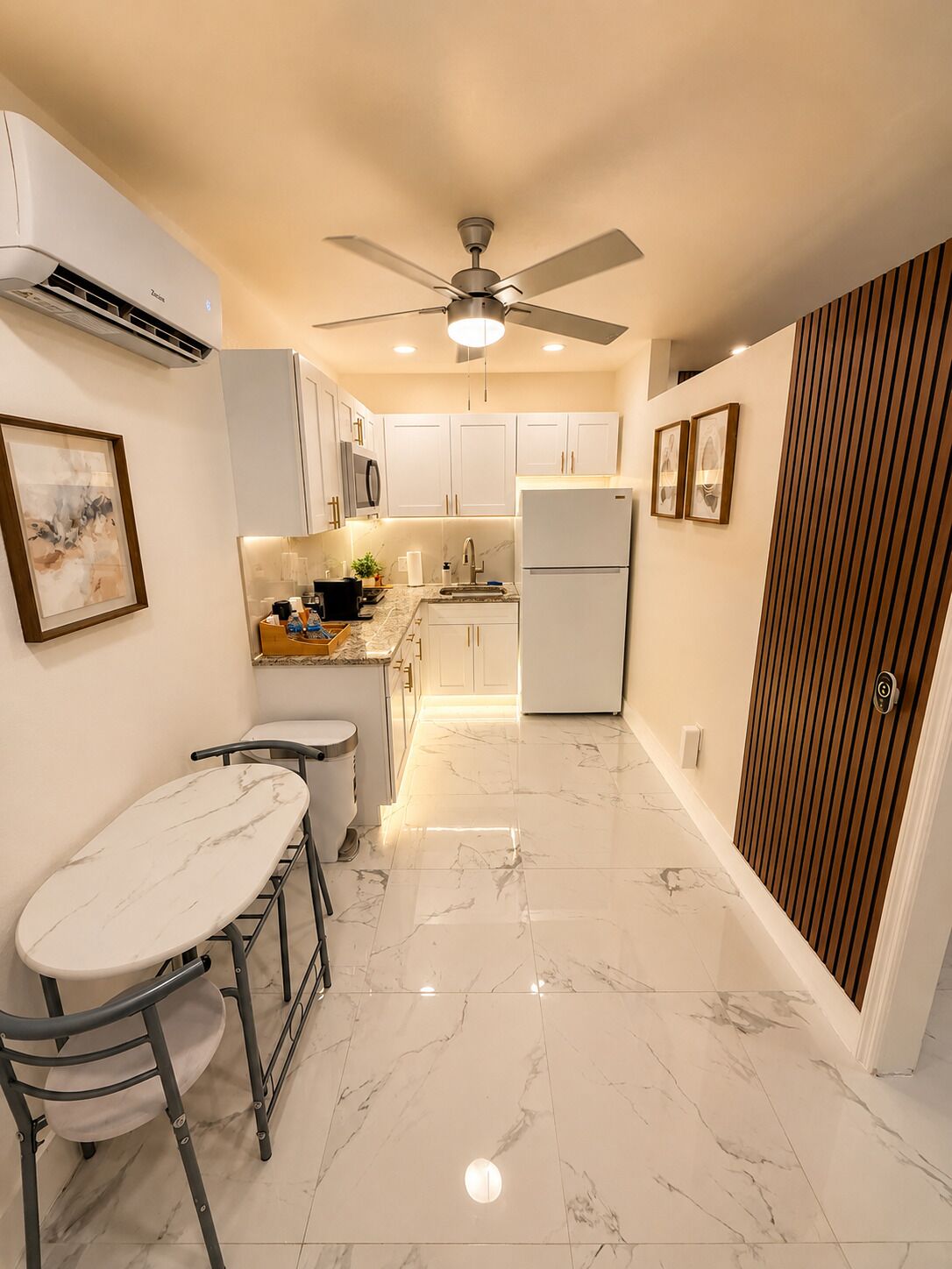 A modern kitchen with white cabinets, a marble countertop, a ceiling fan, and a refrigerator.
