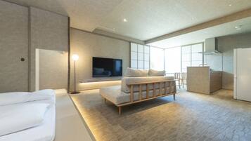 A modern hotel room with a flat-screen TV, a sofa, a bed, a desk, and a kitchen area.