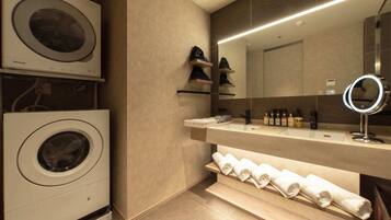 A modern bathroom with a built-in washing machine, a vanity with a mirror, and shelves for towels and toiletries.