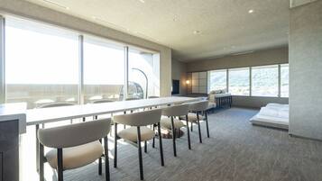 A modern hotel room with a large dining table, multiple chairs, a bed, and large windows offering a view of the outdoors.
