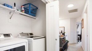 A laundry area with a washing machine and dryer, a shelf with cleaning supplies, and a hallway leading to a room with a mirror and a plant. - Family friendly townhome + Game Room + King beds (Salem)
