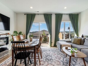 Un saló modern amb zona de menjador, finestres grans amb cortines verdes, un televisor de pantalla plana i vista a una zona residencial.