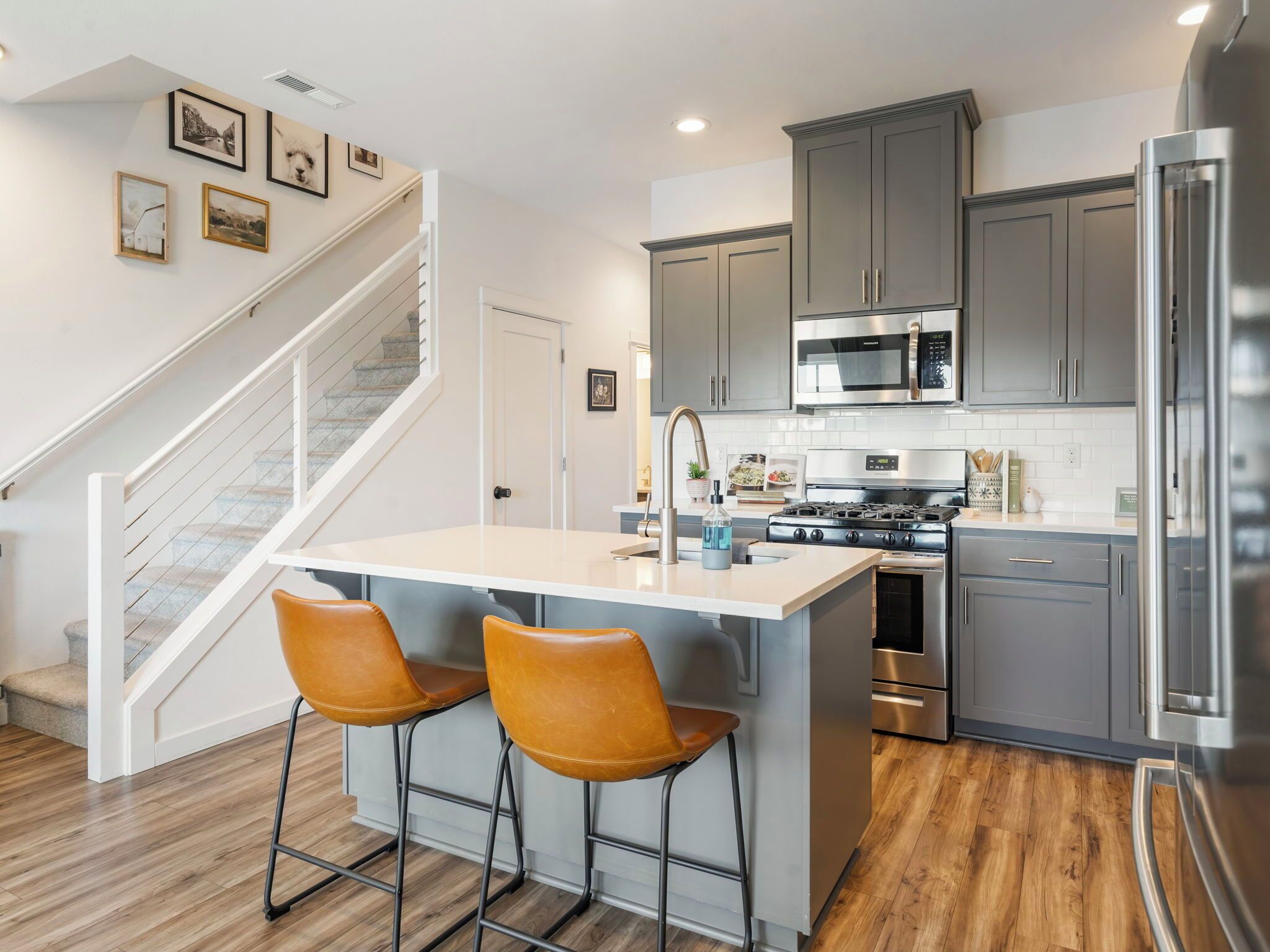 A modern kitchen with grey cabinets, stainless steel appliances, a central island with two chairs, and a wooden staircase.