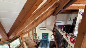 Un dormitorio tipo loft con techo inclinado, vigas de madera, cama con ropa de cama estampada, una mesita y una ventana con vistas a la vegetación.