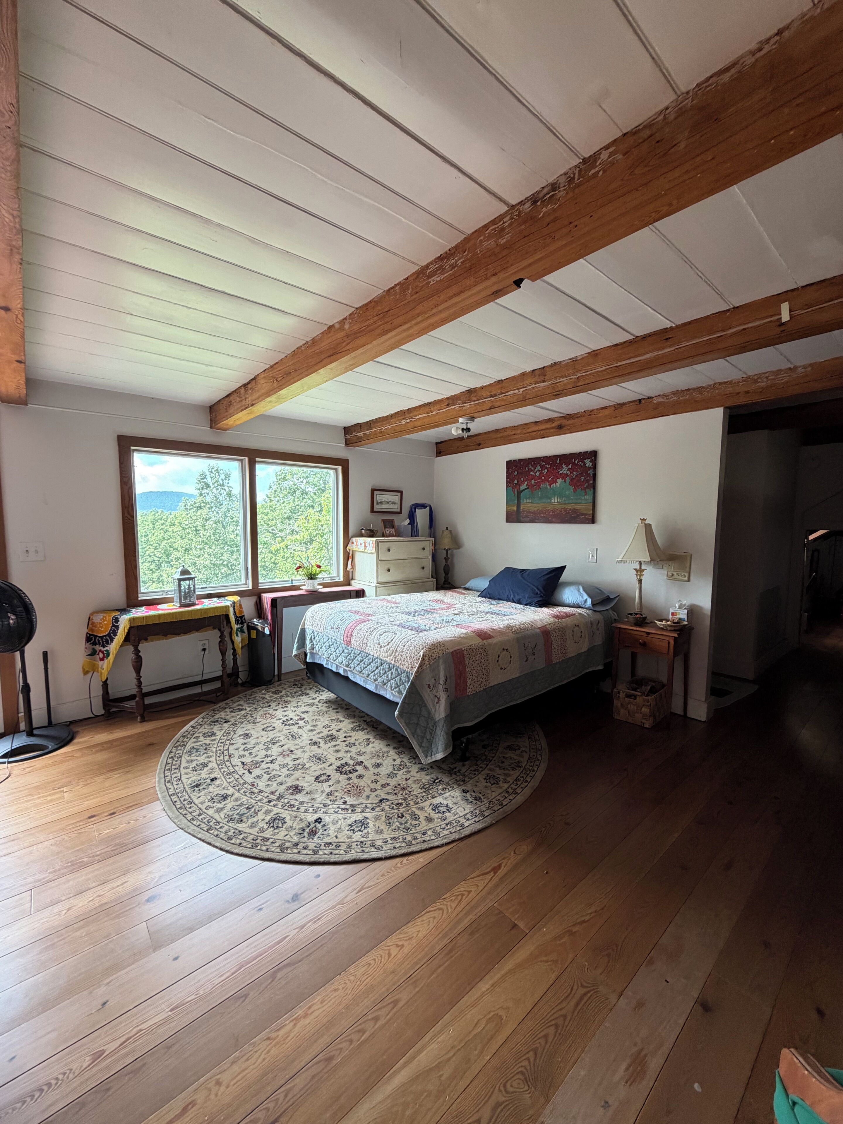 Un dormitorio con techo de madera, una cama con colcha, una mesita de noche, una mesa con lámpara y una ventana con vistas a árboles.