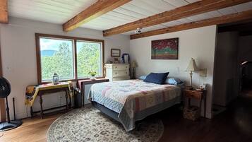 Un dormitorio con techo de madera, una cama con colcha, una mesita de noche, una mesa con lámpara y una ventana con vistas a árboles.