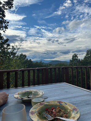 A dining table with plates, cutlery, and a glass on a wooden deck with a view of the sky and trees.