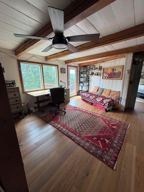 A room with a ceiling fan, wooden beams, a desk, a bed with patterned bedding, and a large rug. - Rustic Cabin with view of Old Rag Mt. 20min to WhiteOak Canyon Shenandoah (Reva)