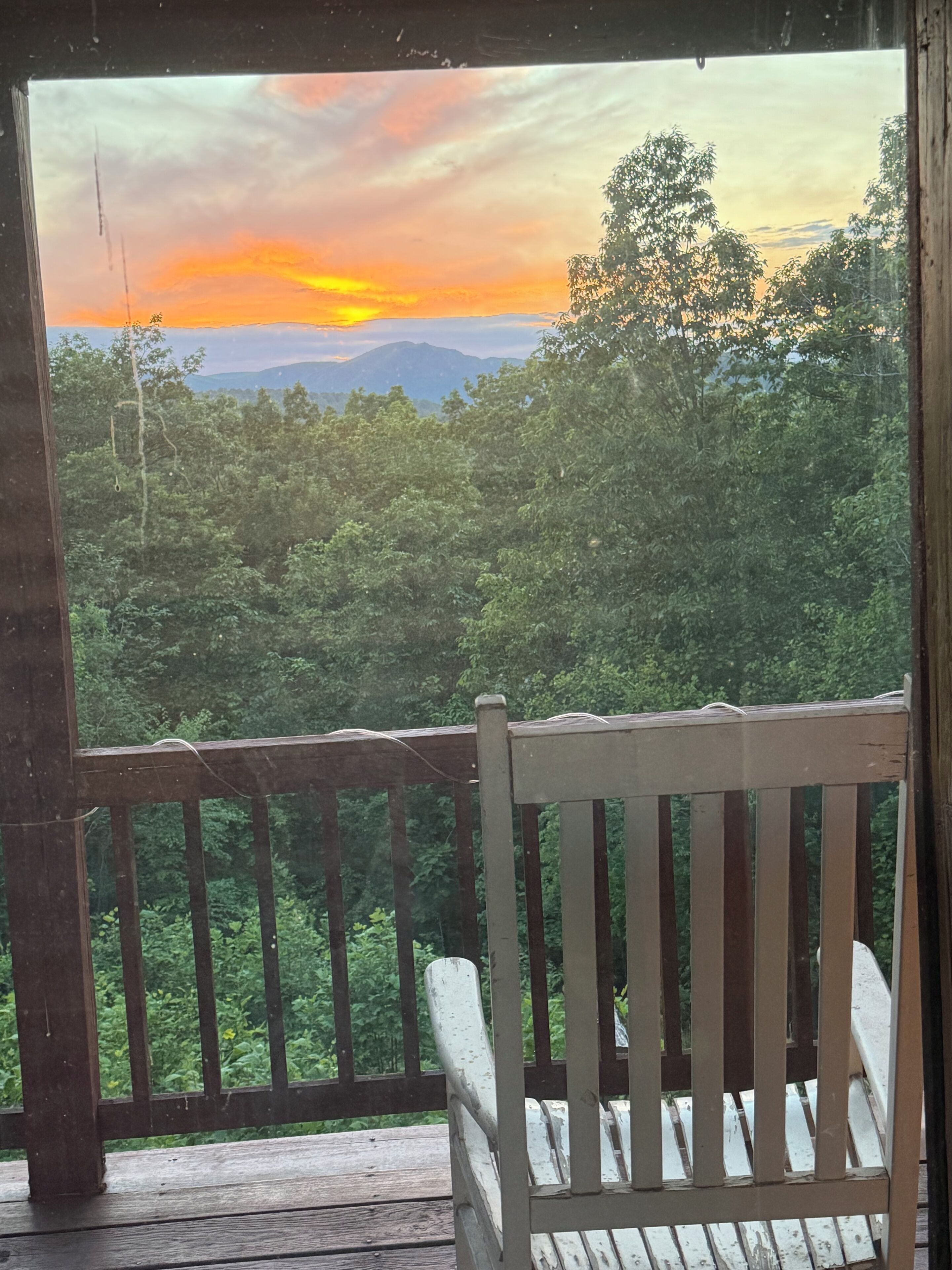 A wooden deck with a white chair, overlooking a forested landscape with mountains in the distance.