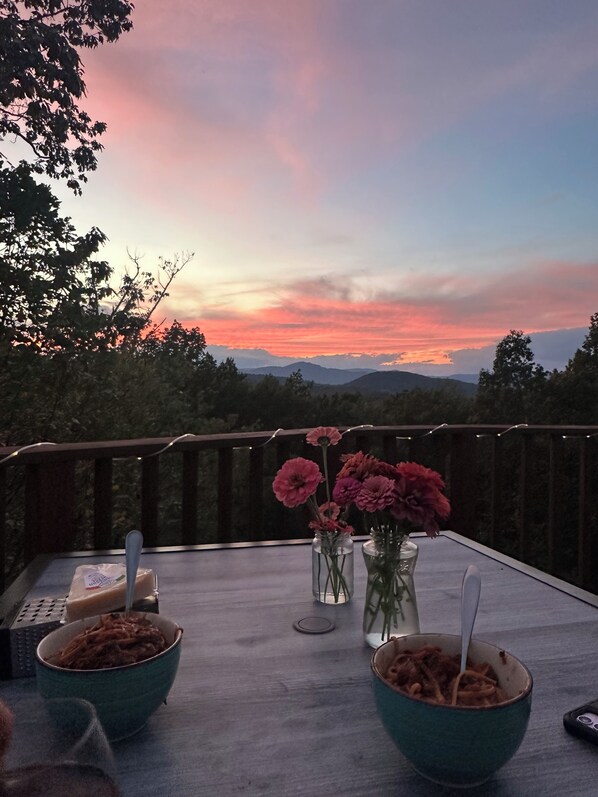 Una mesa con fuentes de comida y jarrones de flores en un balcón con vistas a montañas y un atardecer.