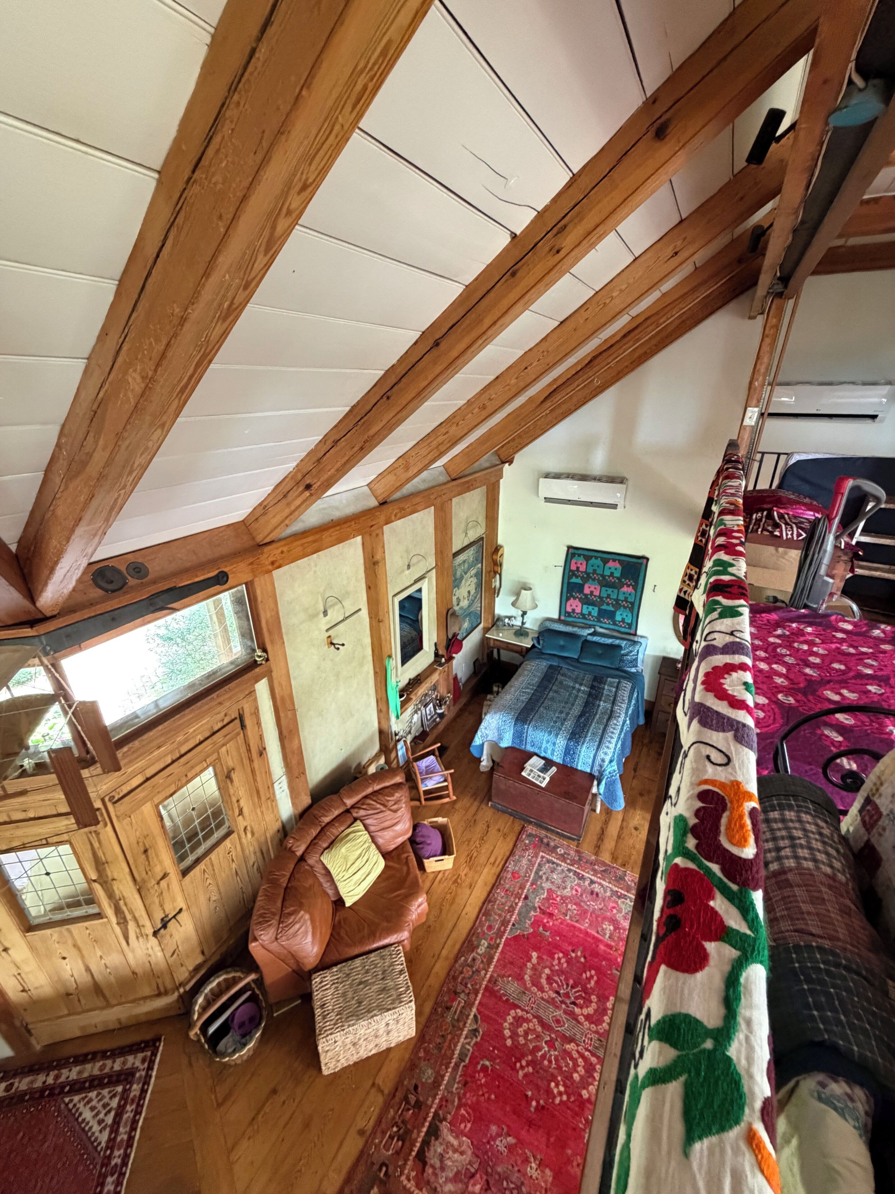 A loft-style bedroom with exposed wooden beams, a bed with a patterned bedspread, a small seating area with a leather chair, and a window providing natural light.