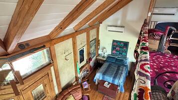 A loft-style bedroom with exposed wooden beams, a bed with a patterned bedspread, a small seating area with a leather chair, and a window providing natural light.