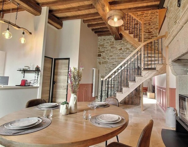 A dining area with a wooden table set for four, a stone wall, and a staircase leading to an upper level. - Le Cerf - LANGAST - Vacation Rental (LANGAST)