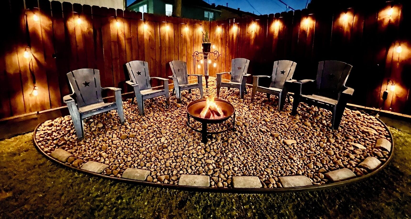 Een rond zitgedeelte buiten met een vuurkorf in het midden, omringd door stoelen en een houten hek met lichtjes.