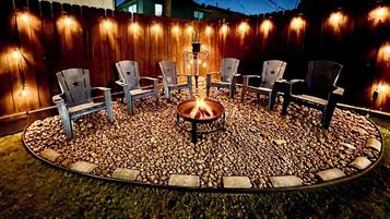 Een rond zitgedeelte buiten met een vuurkorf in het midden, omringd door stoelen en een houten hek met lichtjes.
