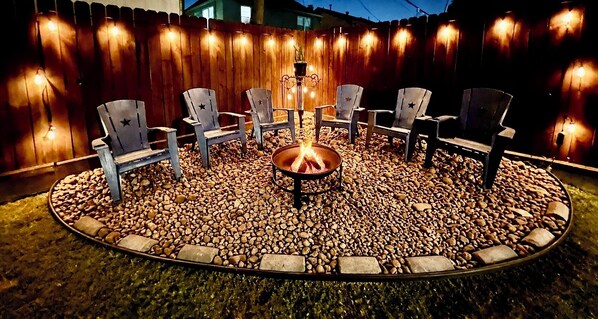 A circular outdoor seating area with a fire pit at the center, surrounded by chairs and a wooden fence with string lights.
