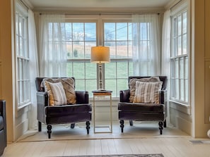 A room with two leather armchairs, a small table, and a lamp. There are large windows with curtains.
