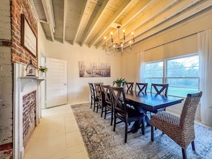 A dining area with a wooden table and chairs, a chandelier, a fireplace, and a window with a view of trees.