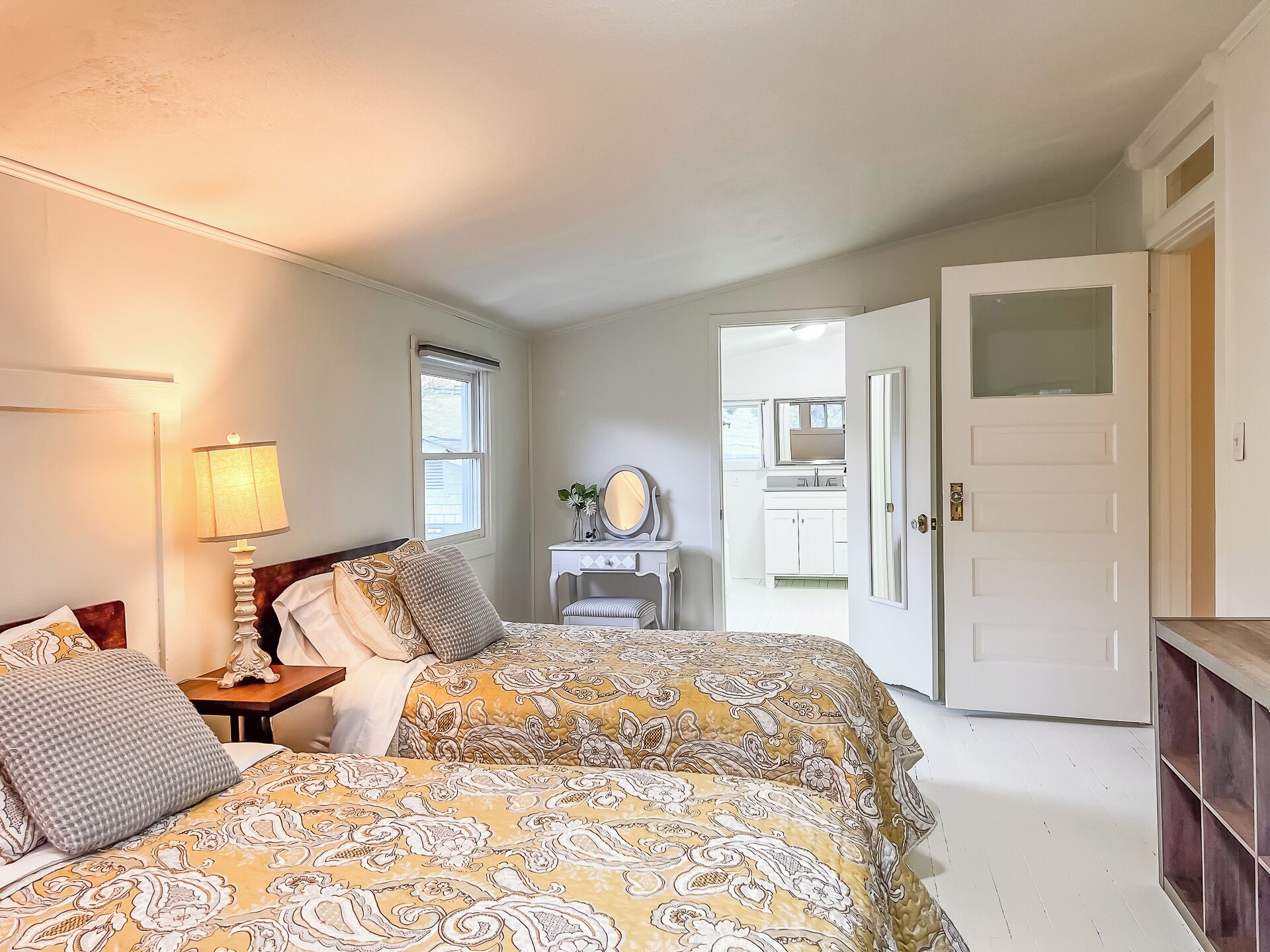 A bedroom with a bed, bedside table, lamp, dresser, mirror, and a door leading to another room.
