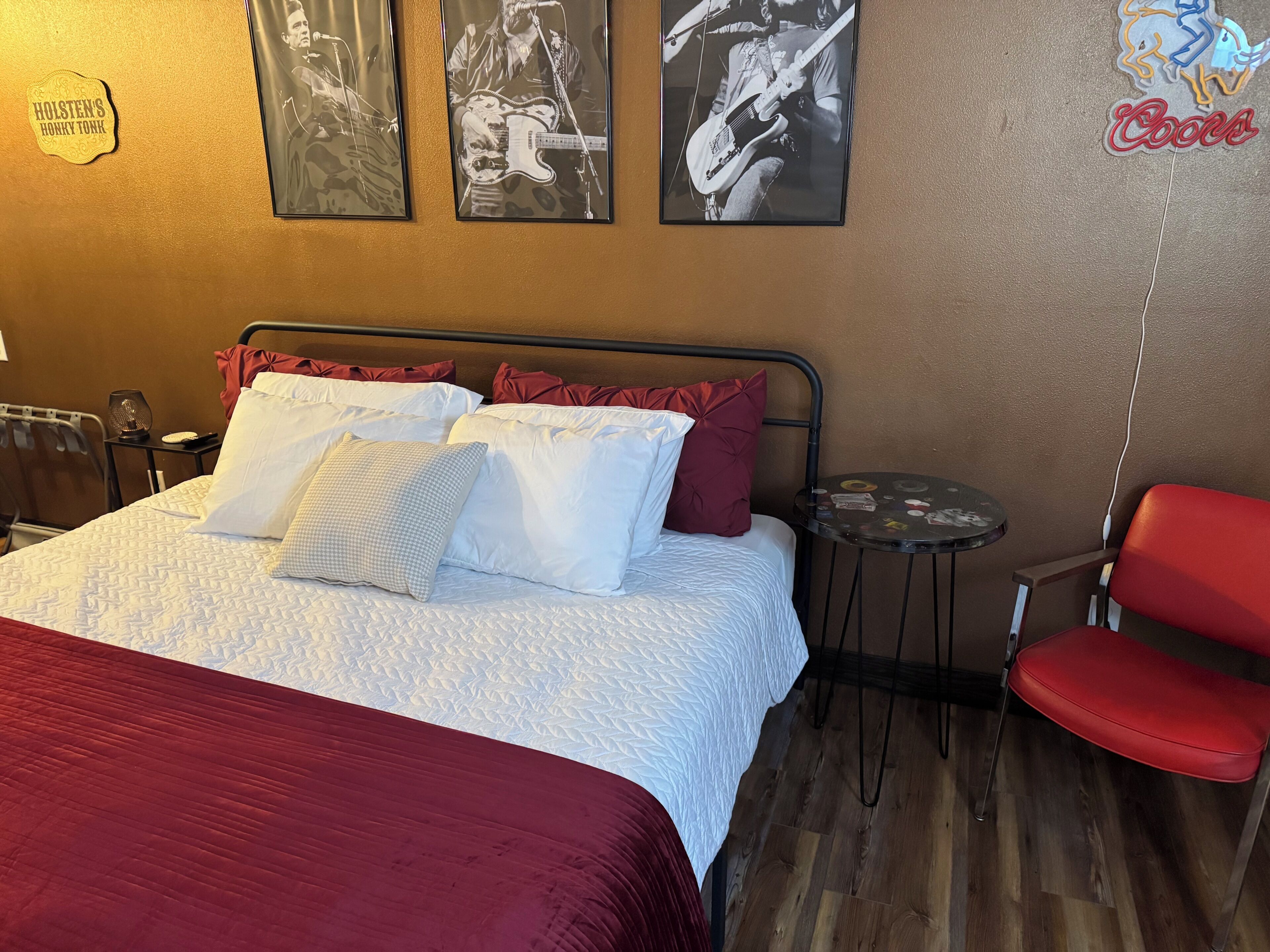 Een hotelkamer met een bed, kussens, een rode stoel, een klein tafeltje en wanddecoraties, waaronder ingelijste foto’s en een neonbord.