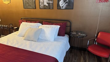Een hotelkamer met een bed, kussens, een rode stoel, een klein tafeltje en wanddecoraties, waaronder ingelijste foto’s en een neonbord.