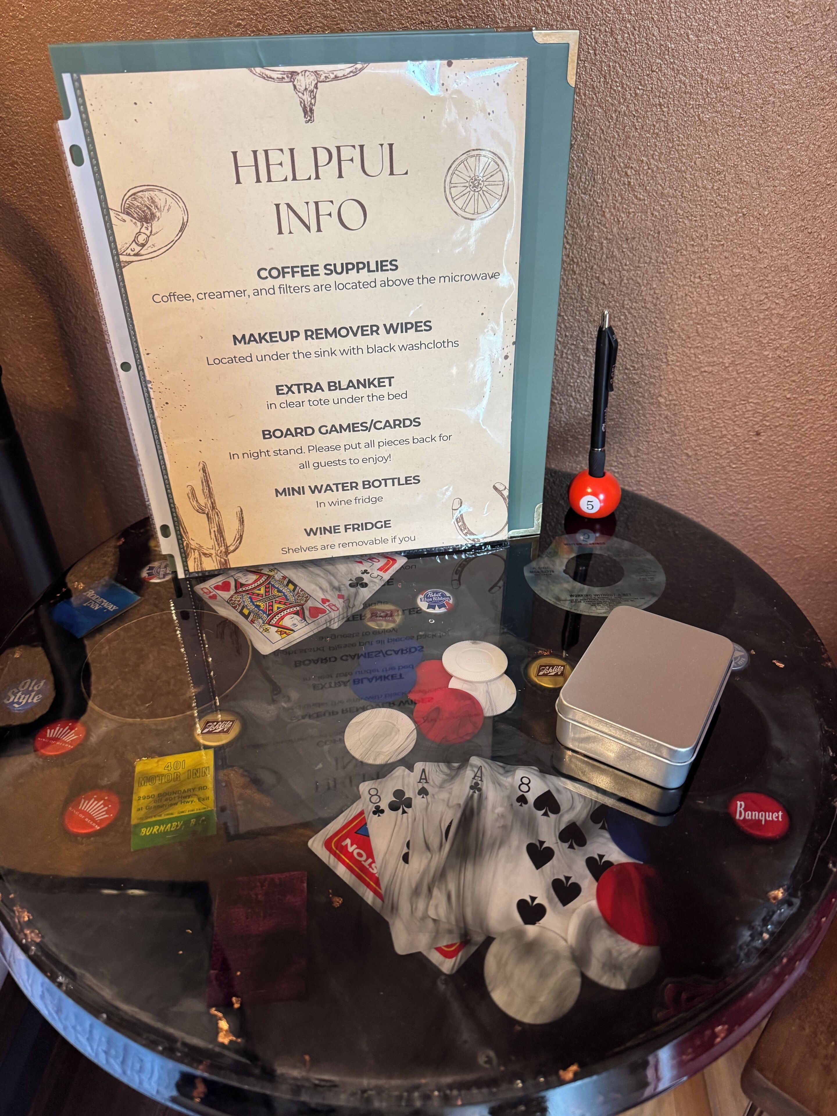 A glass table with a sign listing helpful info for coffee supplies, makeup remover wipes, an extra blanket, board games/cards, mini water bottles, and a wine fridge.