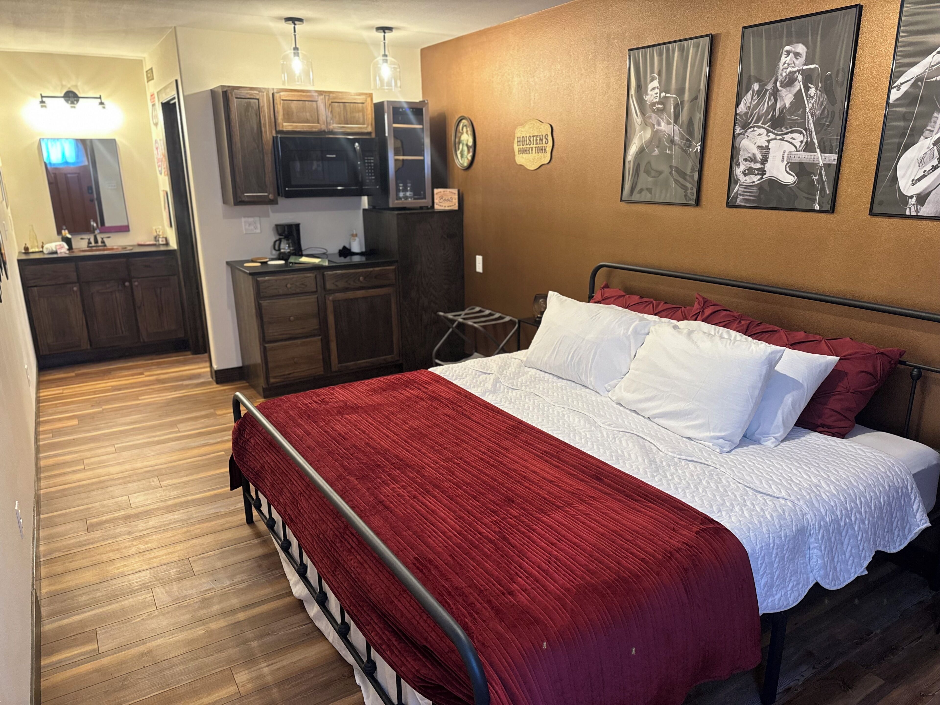 A bedroom with a bed, a red blanket, and a white pillow. There is a kitchenette with a microwave and cabinets.