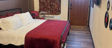 A bedroom with a bed, a red blanket, a ceiling fan, a window with curtains, and a door.