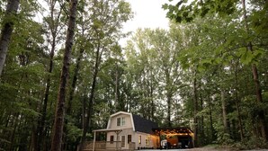 A cabin in a forest with a gravel driveway and a covered outdoor seating area.