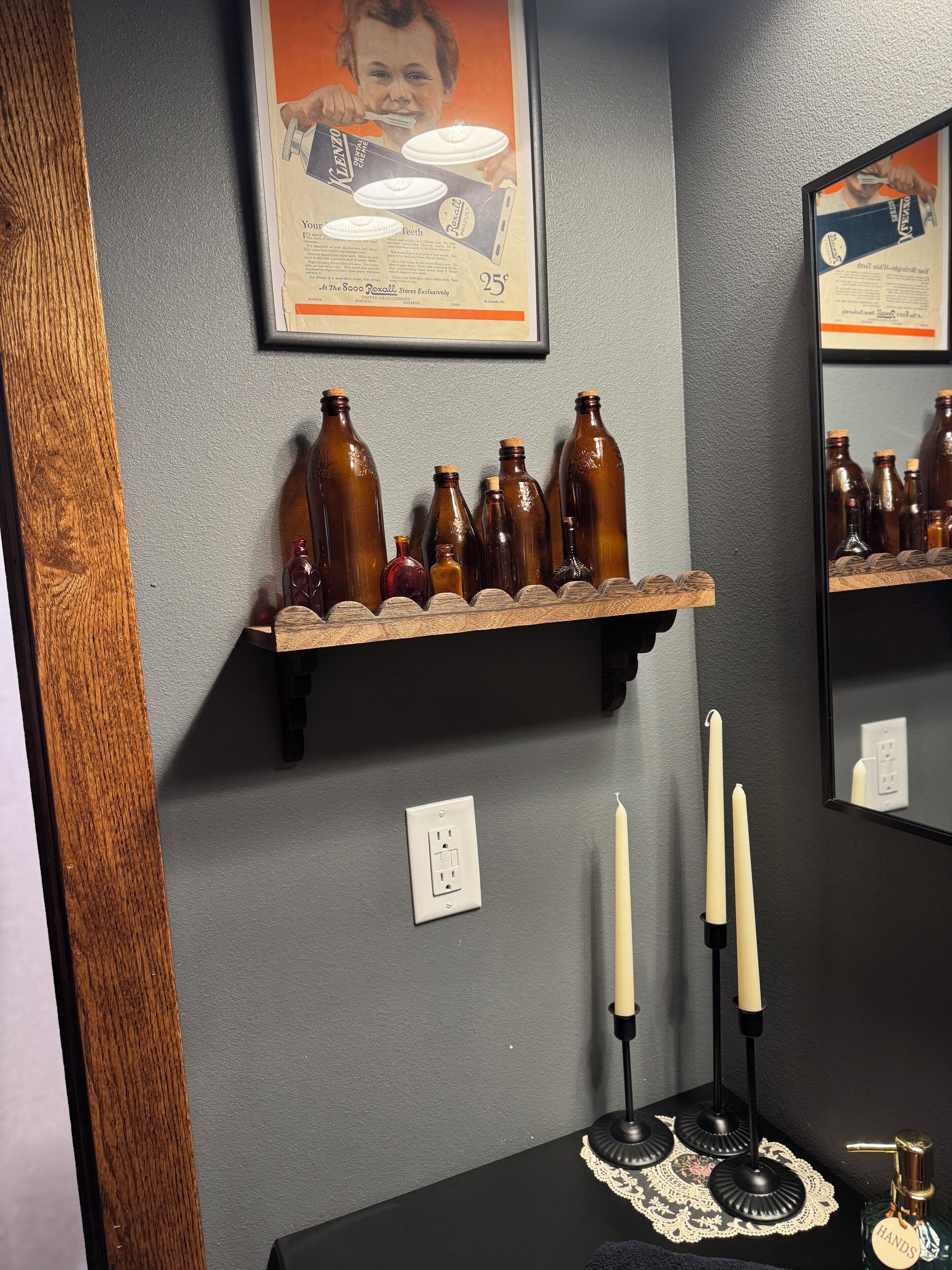 A bathroom with a wooden shelf holding brown bottles, a framed advertisement, a mirror, and a black countertop with candles.