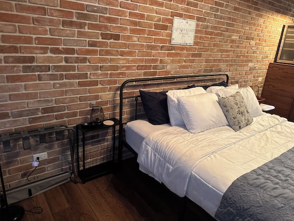 Una habitación con pared de ladrillo, estructura de cama metálica, ropa de cama blanca y una mesita de noche con lámpara.
