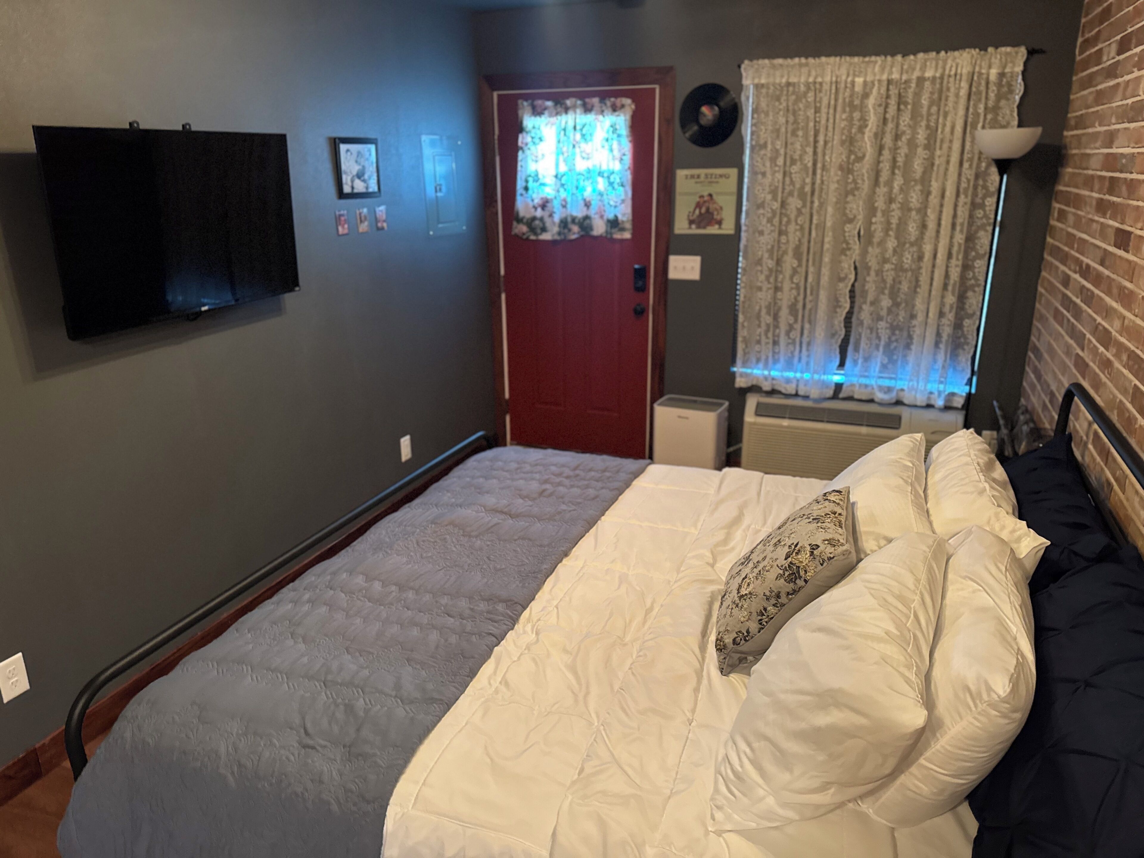 A bedroom with a bed, a flat-screen TV mounted on the wall, a door with a floral curtain, and a wall-mounted air conditioner.