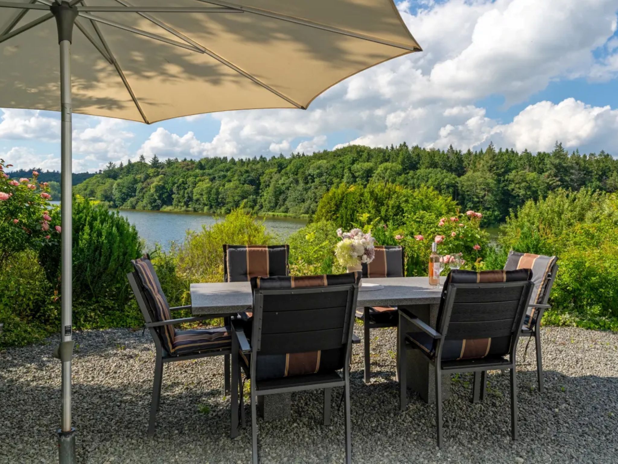 Ein Außenbereich zum Essen mit Tisch und Stühlen, einem großen Sonnenschirm und einem schönen Blick auf einen See und Wald.