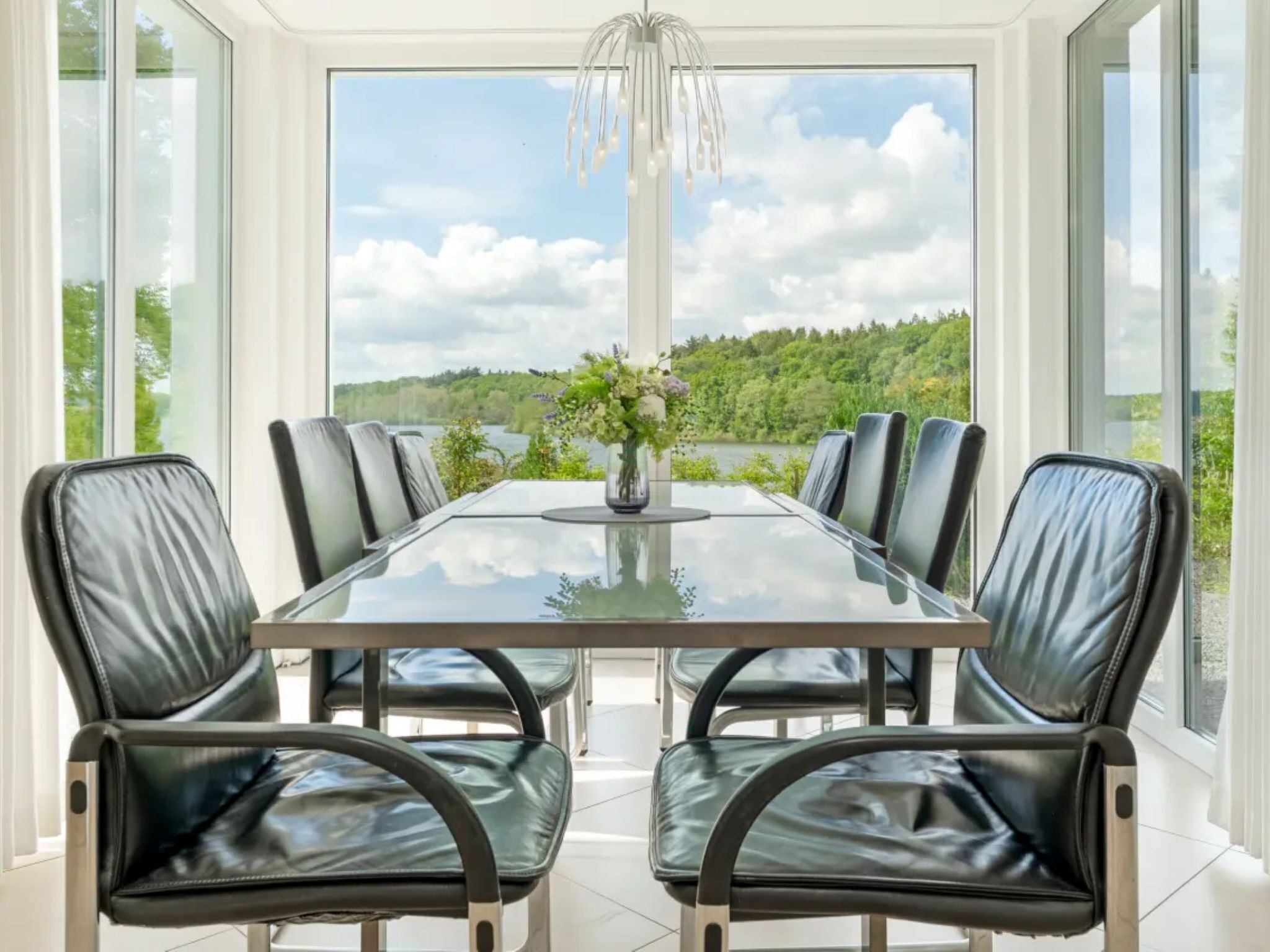 A dining area with a glass table and black leather chairs, a vase with flowers, and a view of a lake and forest through large windows.