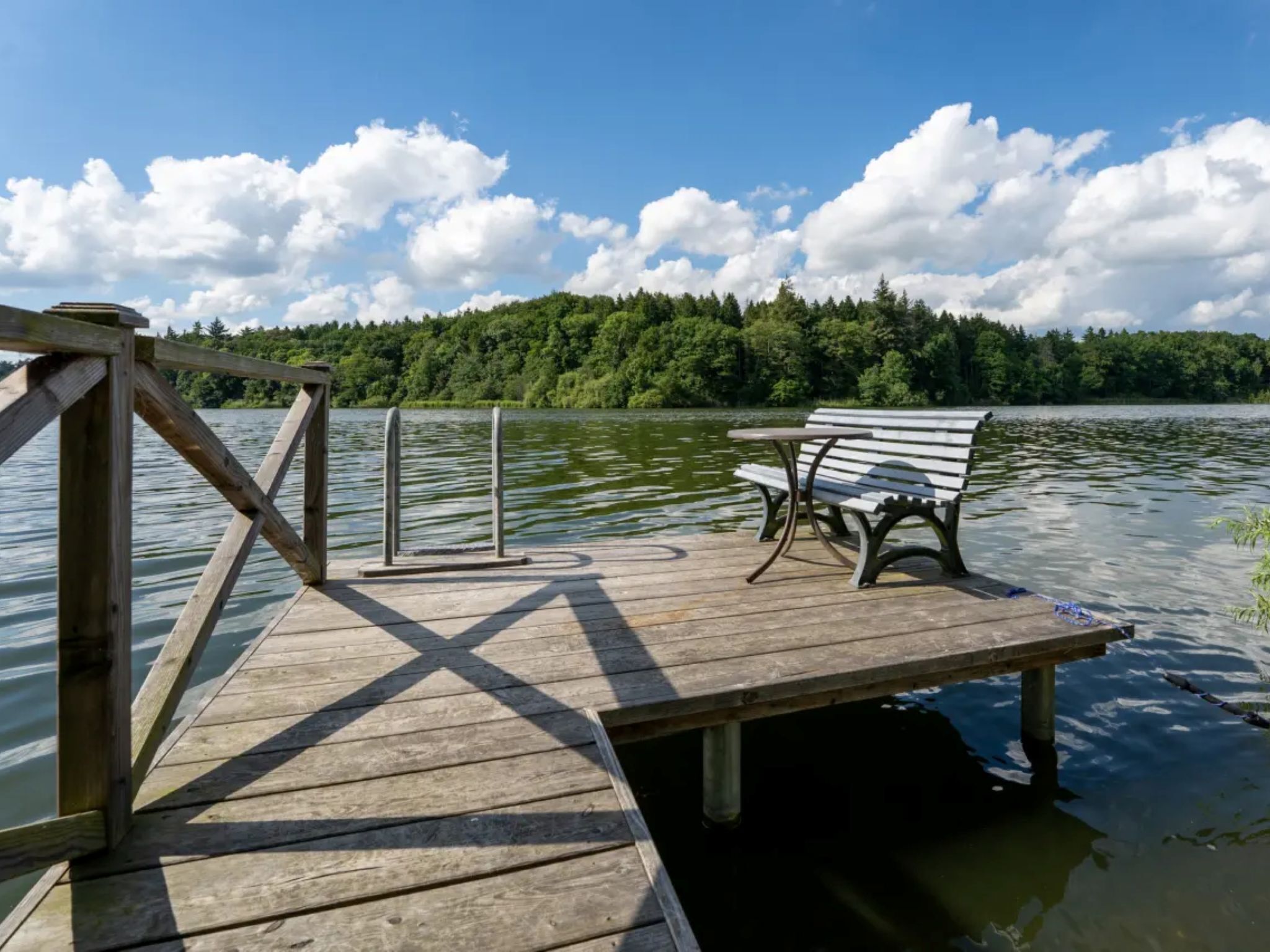 A wooden dock extending into a lake with a bench and a table, surrounded by a forested area.