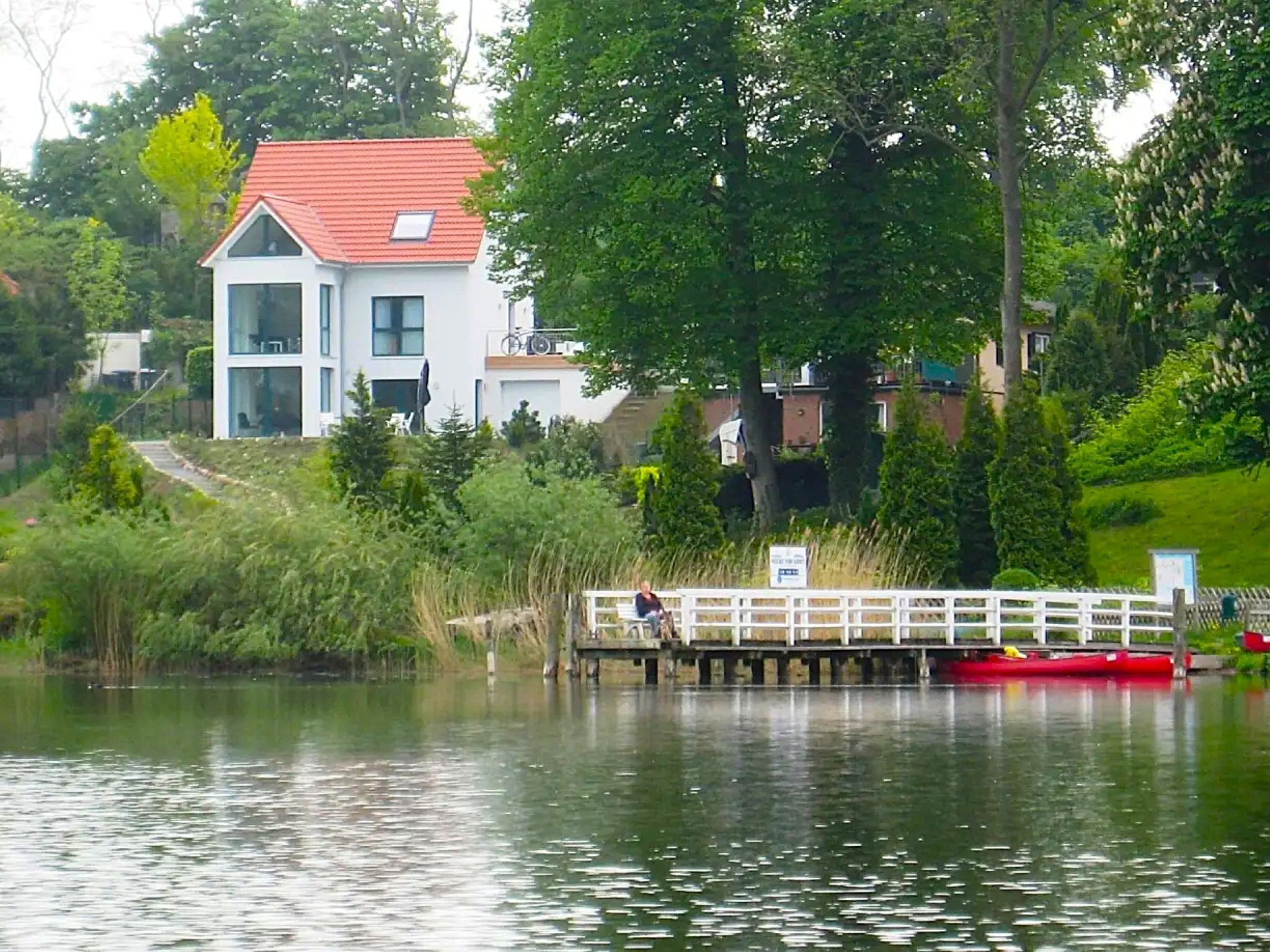A house with a red roof, a dock extending into a body of water, and a person sitting on the dock.